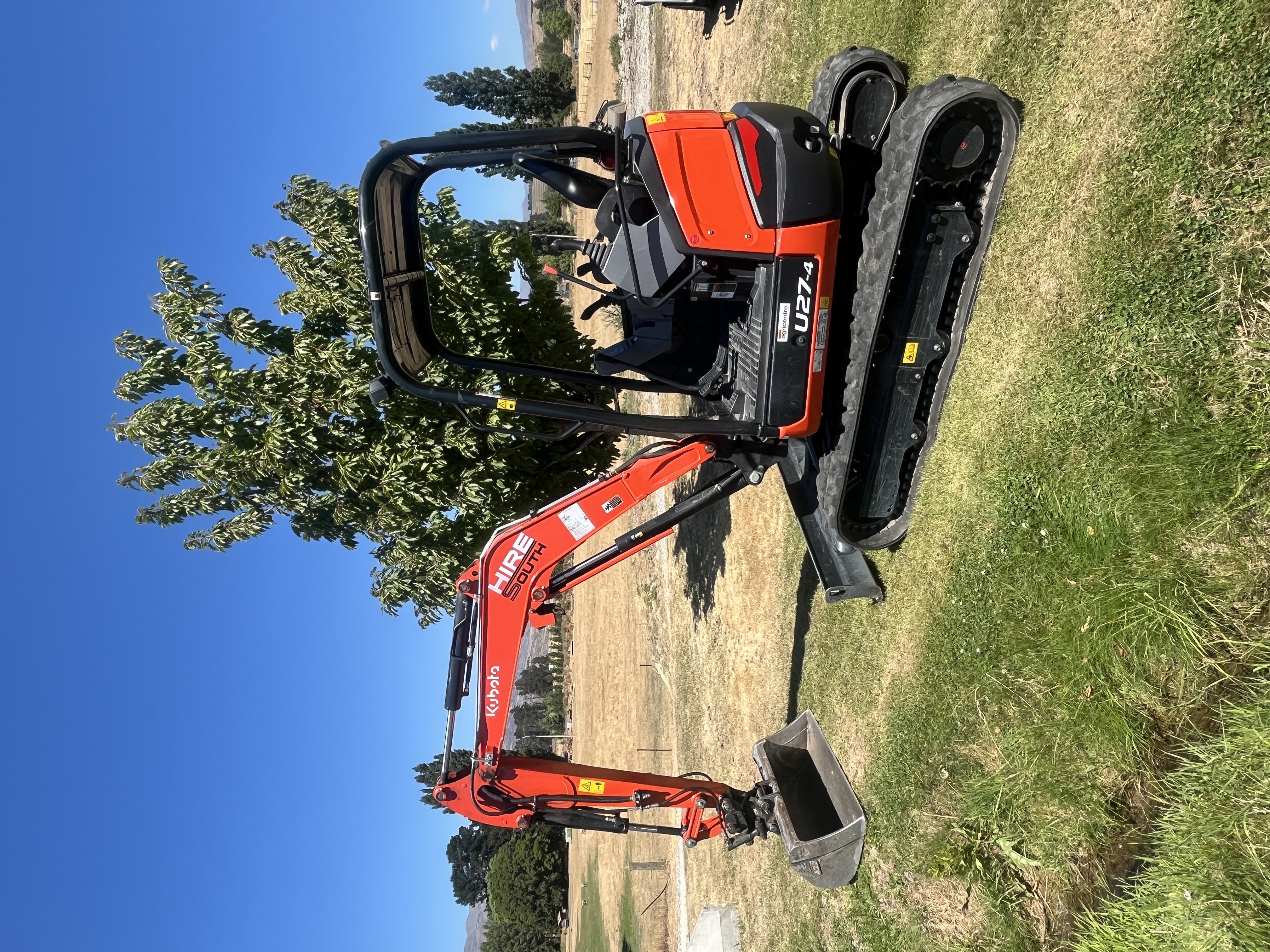 Hire South branded Kubota U27-4 digger parked in a paddock, clean side profile showing Hire South livery and Central Otago mountains in the background