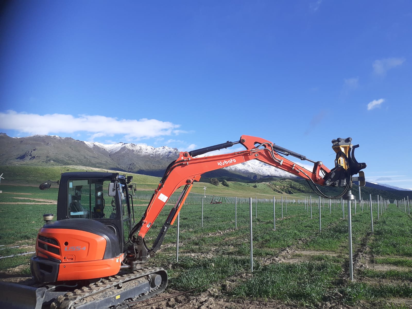 Hire South Kubota U17 mini digger with post driver attachment working a vineyard row, Pisa Range snow-capped in the background