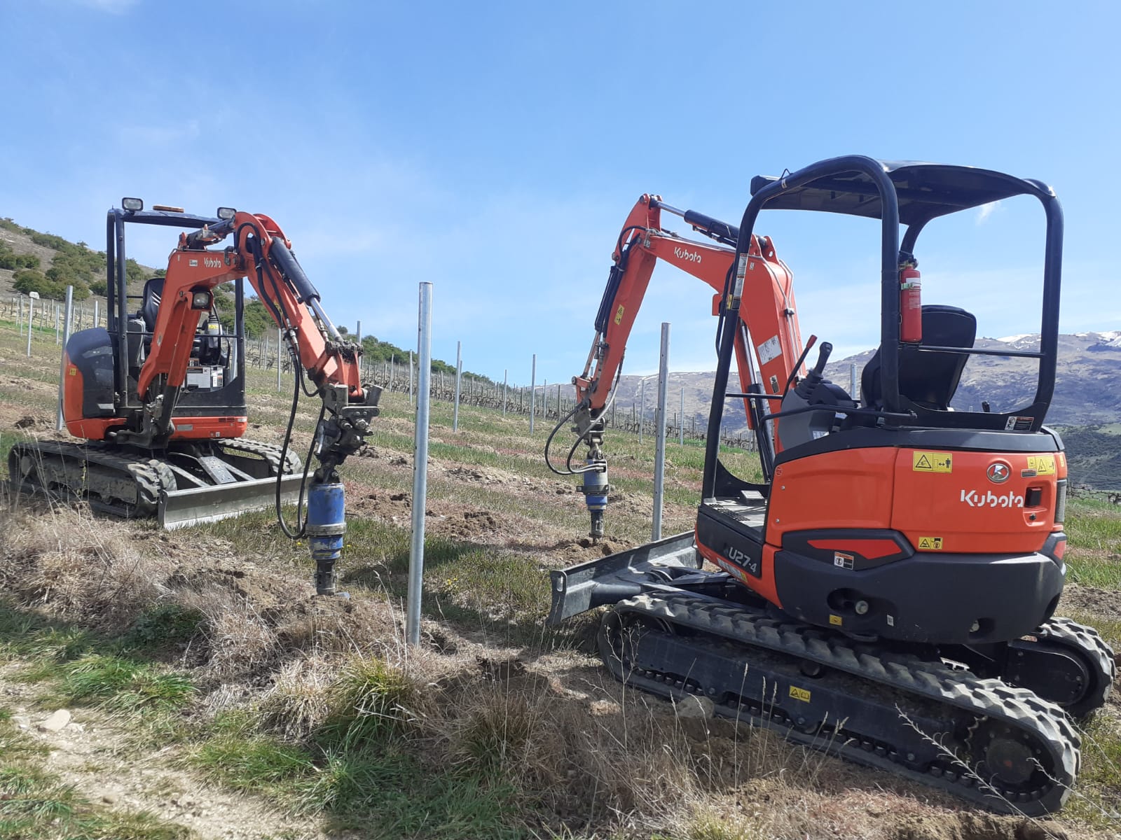 Two Hire South Kubota U27-4 diggers working alongside each other driving posts in a Central Otago vineyard, snowy ranges behind