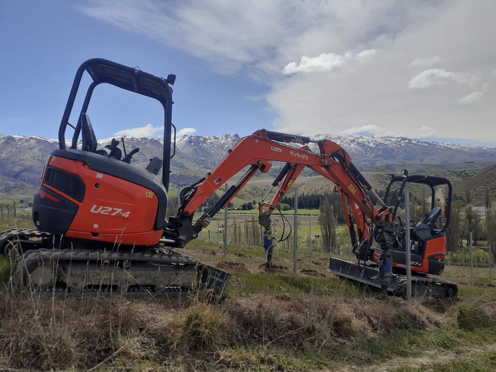 Hire South Kubota U27-4 digger with post driver attachment, set against a sweeping Central Otago valley and snow-capped mountain backdrop