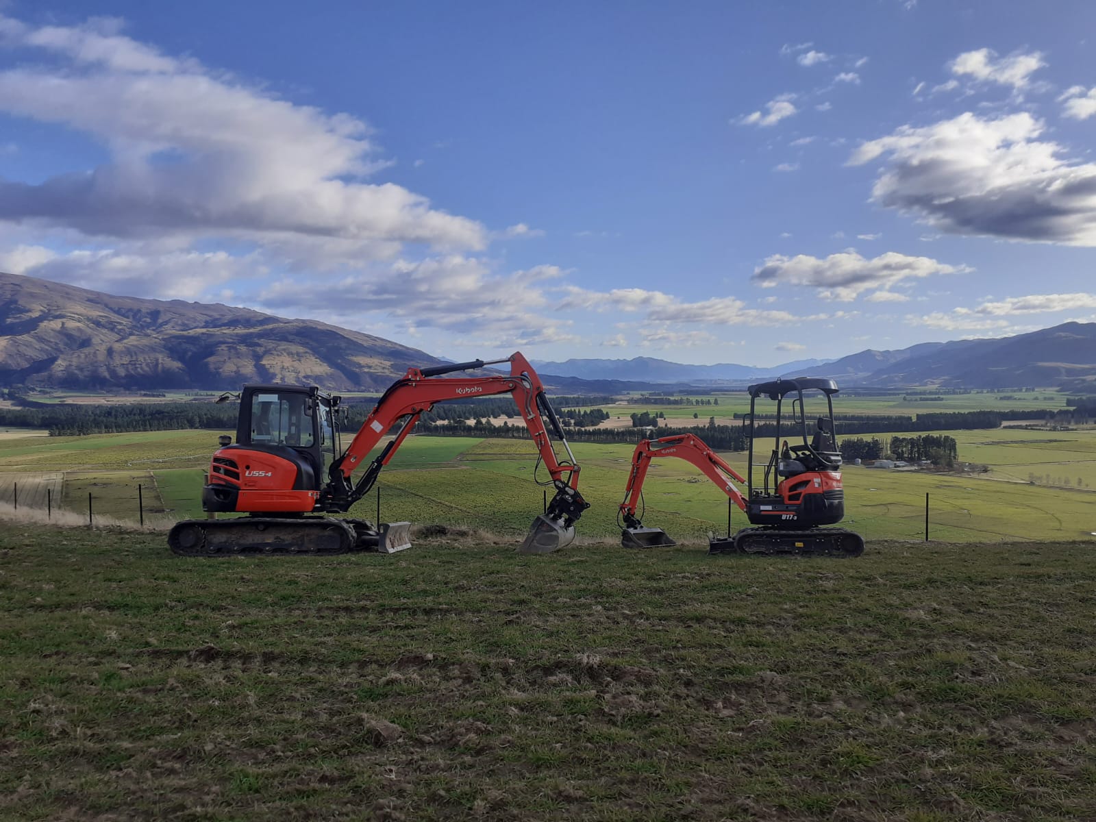 Hire South Kubota U55-4 and U17-3 mini diggers parked side by side in a paddock with a wide panoramic view of the Central Otago valley, mountains, and farmland stretching to the horizon