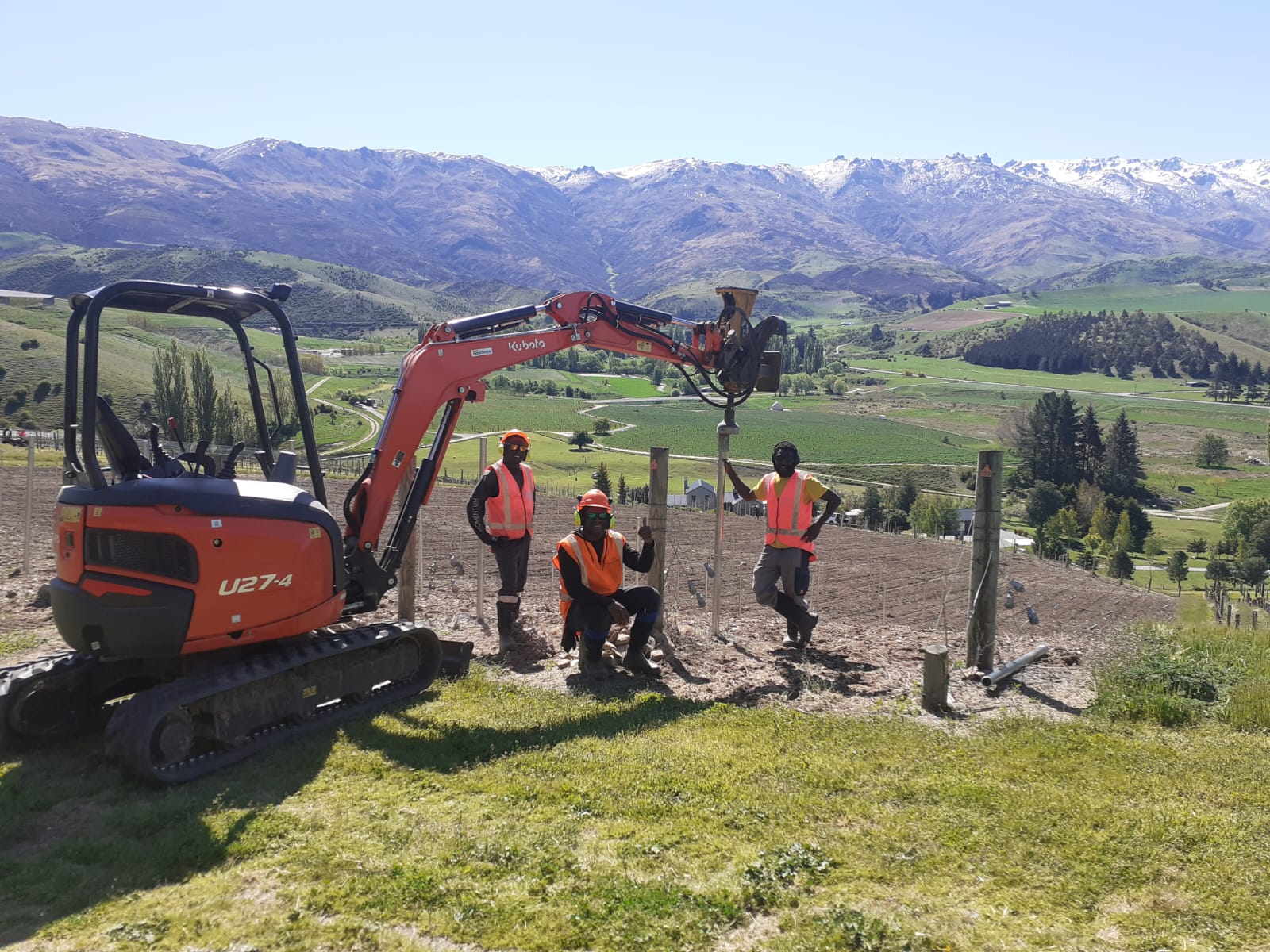 Hire South team of three workers with a Kubota U27-4 digger after completing a vineyard fence posting job in Central Otago, snow-capped mountains in the background