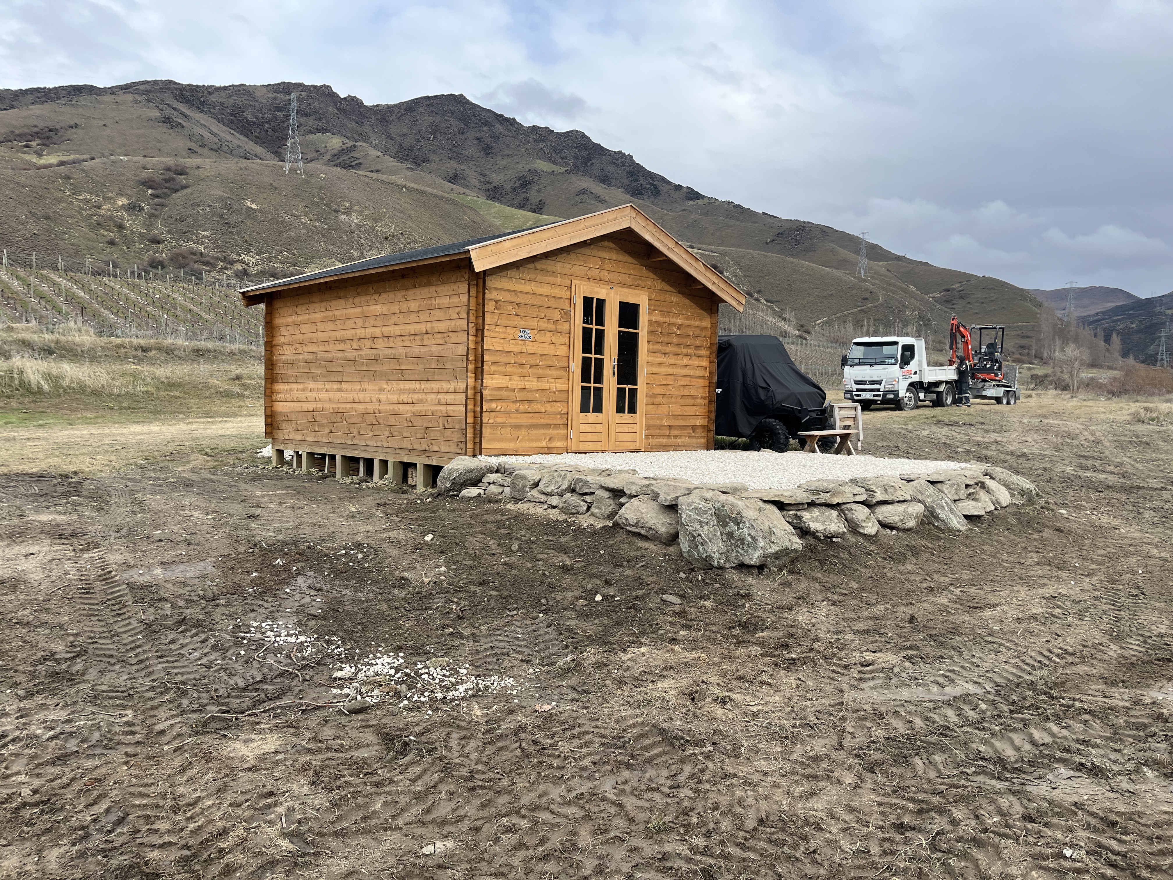 Completed timber cabin with a dry-stone retaining wall and gravel pad, built as part of a Hire South contracting project on a Central Otago hillside with mountains behind