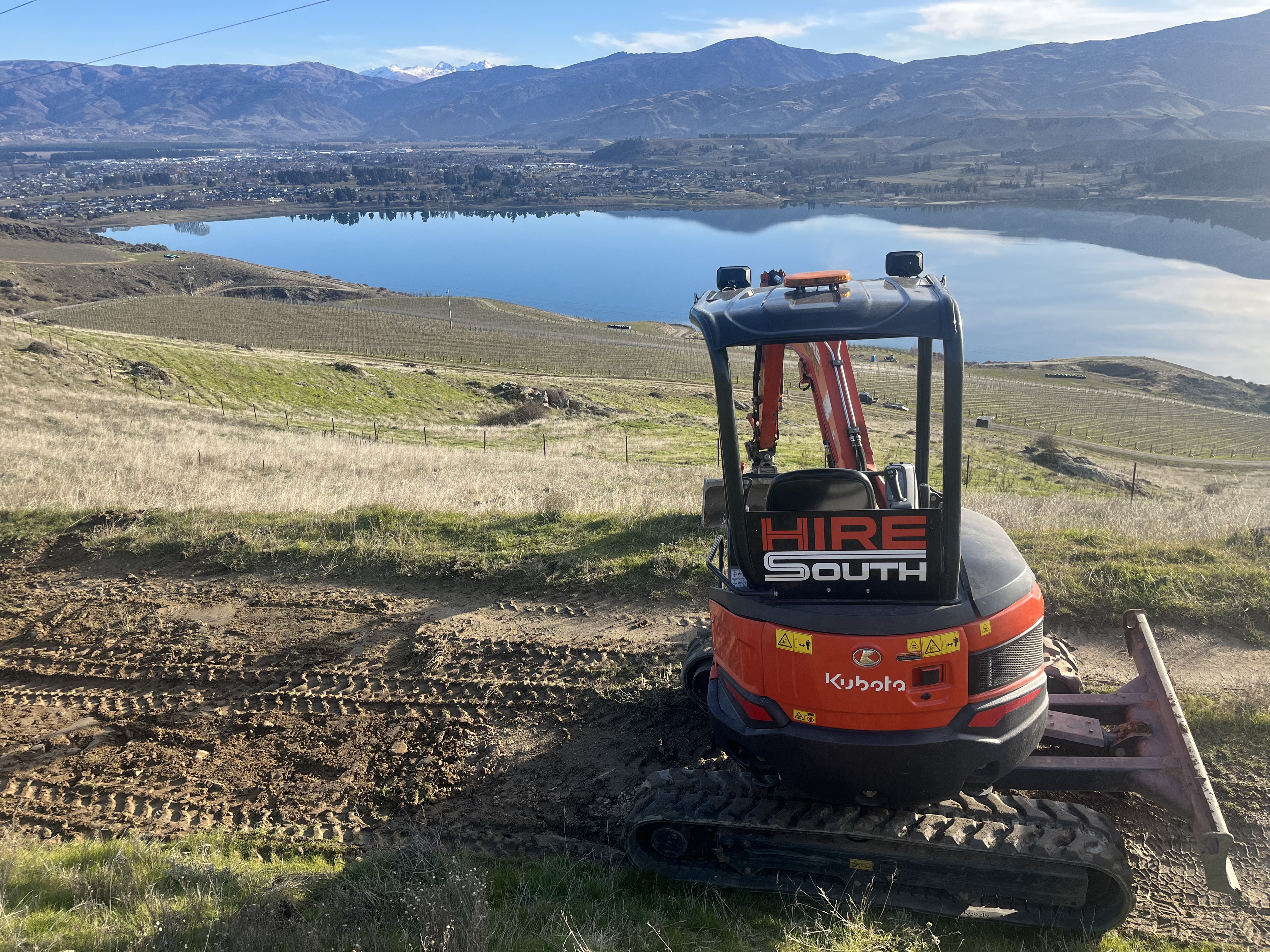 Rear of a Hire South branded Kubota U27 digger with Lake Dunstan and the town of Cromwell perfectly reflected in the calm water below, Central Otago