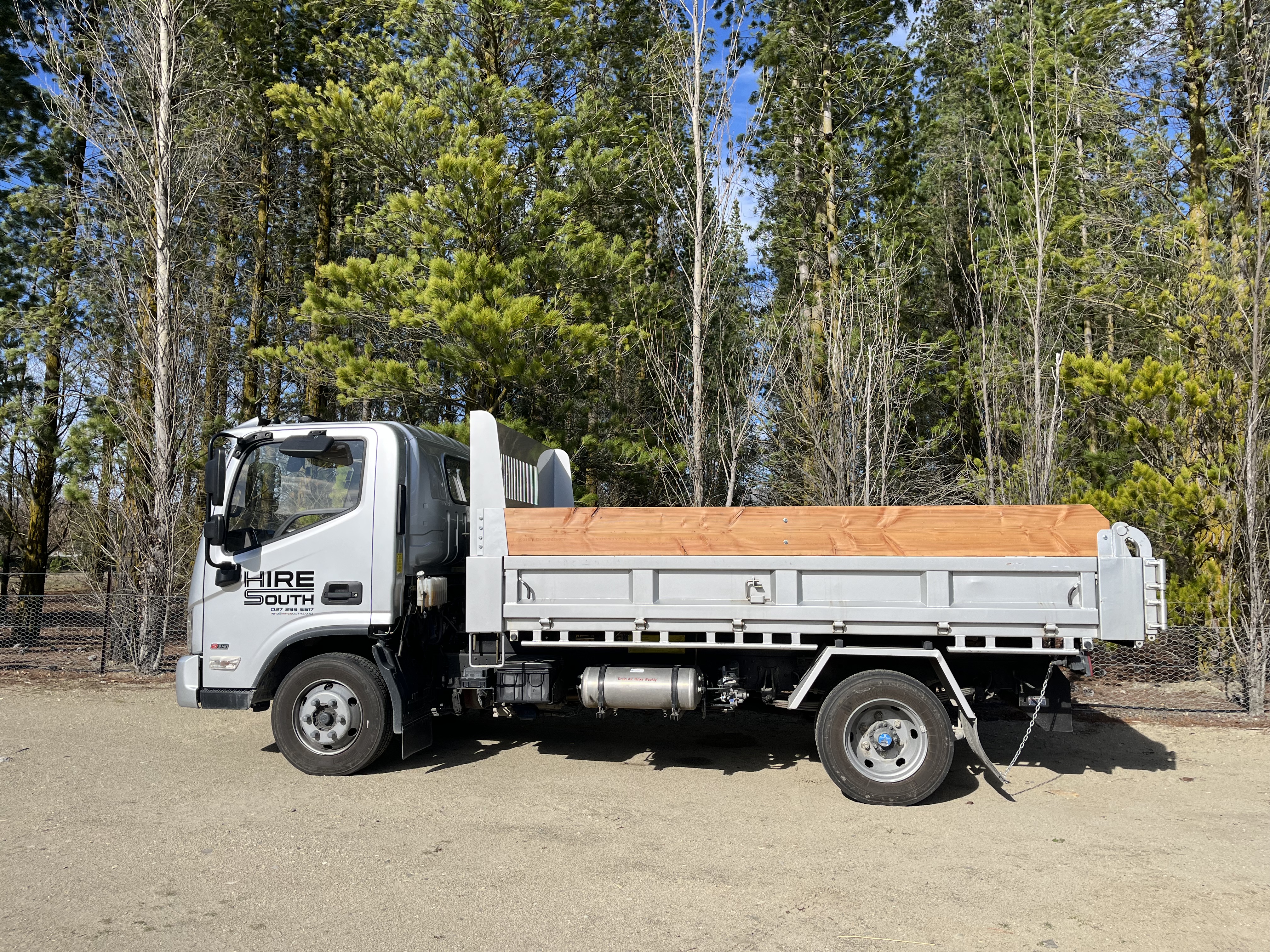 Hire South branded white flatdeck truck with timber deck, side profile, parked in front of trees in Central Otago
