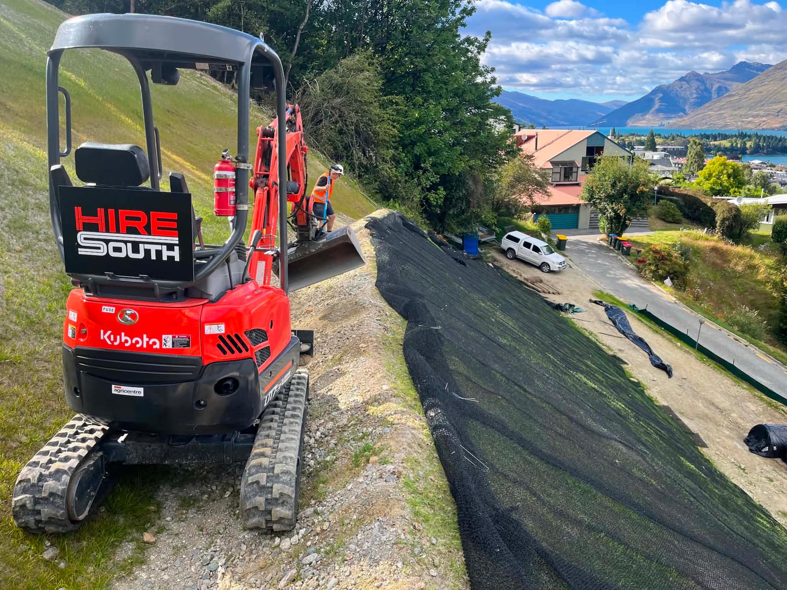 Hire South branded Kubota mini digger with operator forming a new driveway on a steep section above Lake Wakatipu and the Queenstown ranges, Central Otago